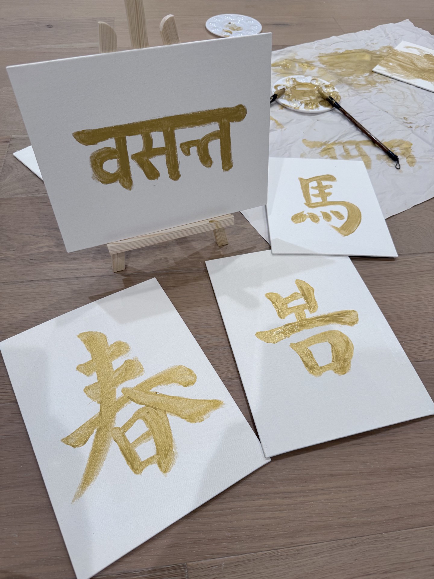 Gold calligraphy on white canvases: 春, 馬, 봄 and वसन्त, with brushes and a palette nearby.