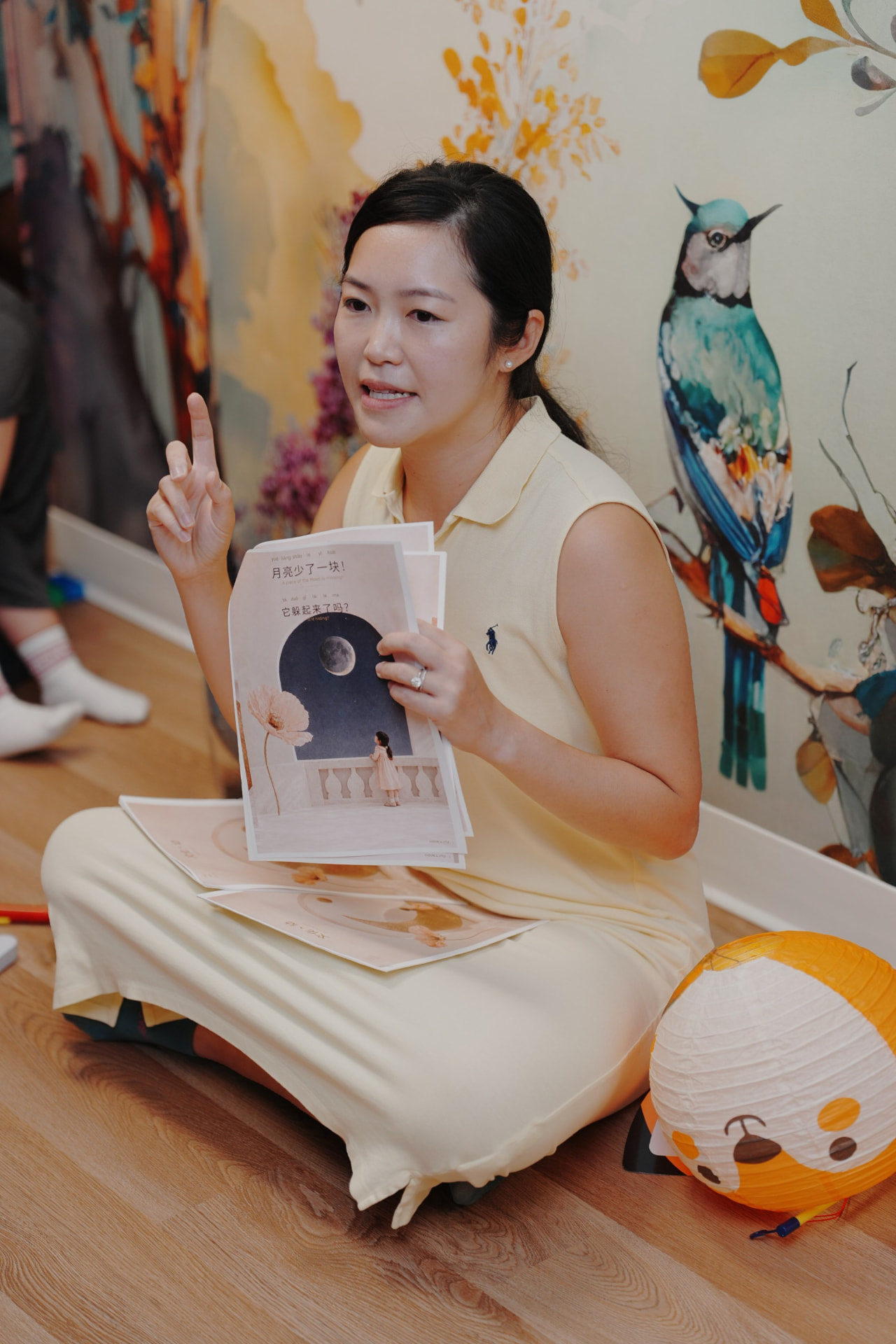 Amanda Lee Liu reading Culture & Little moon pages aloud to a group, seated on the floor beside a paper lantern.